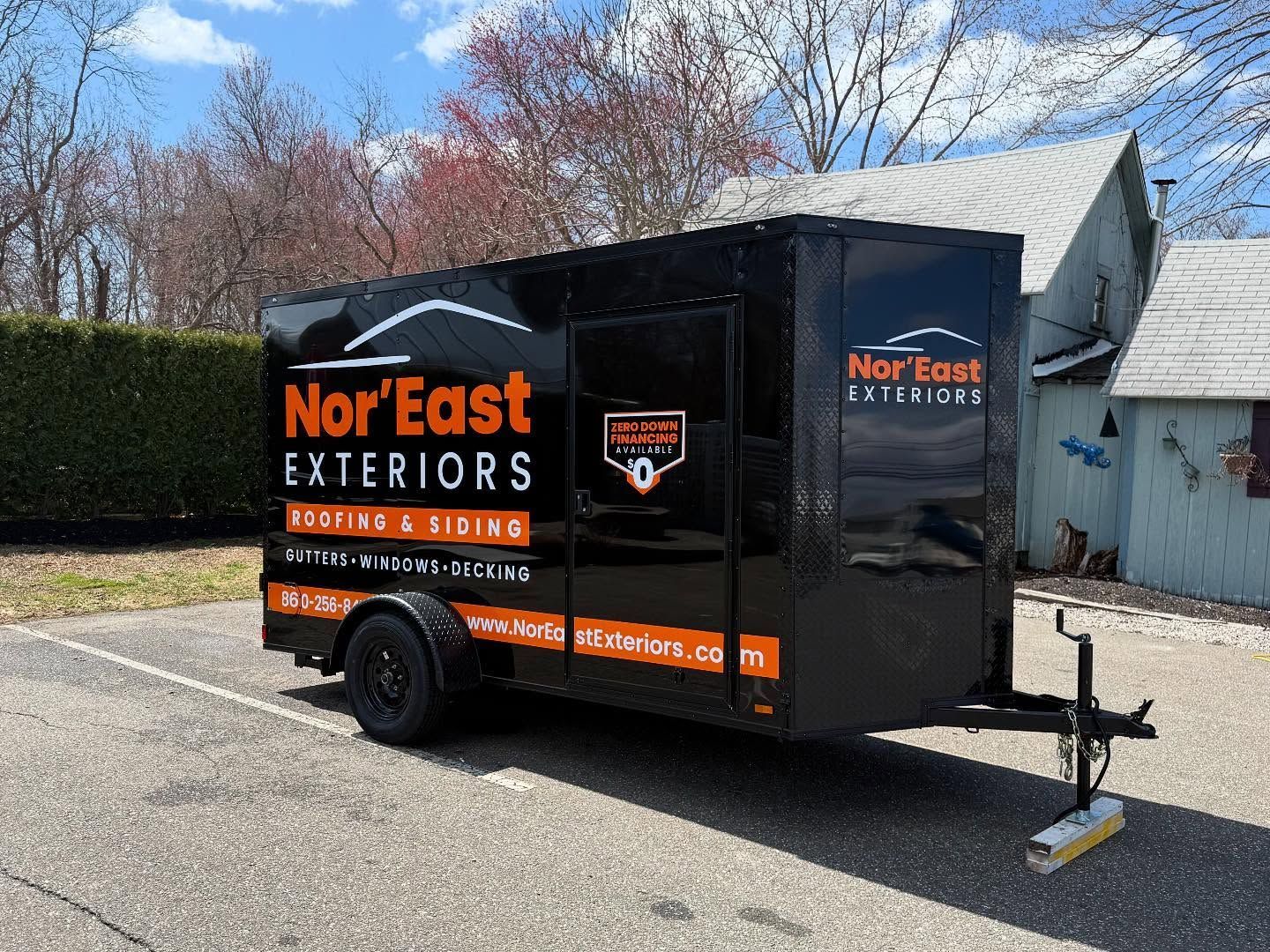 Black Nor'East Exteriors trailer with orange logo, parked on pavement. Building and trees in the background.