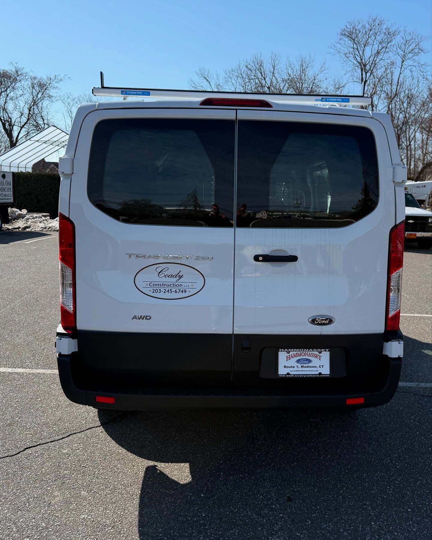 White Ford Transit van, rear view, parked outdoors. Has a roof rack and logo on the back.