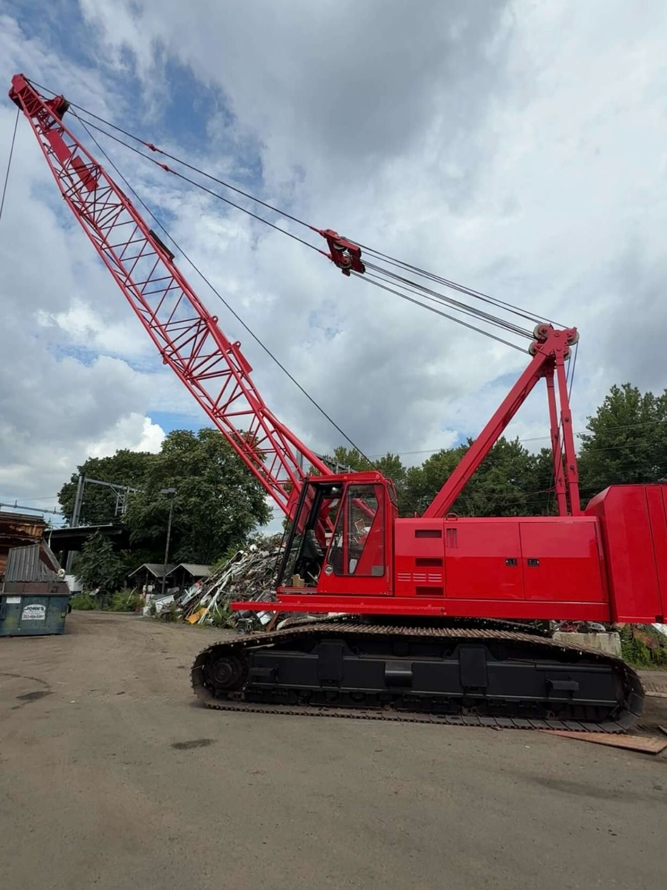 Red crawler crane on a paved surface with a tall boom against a cloudy sky.