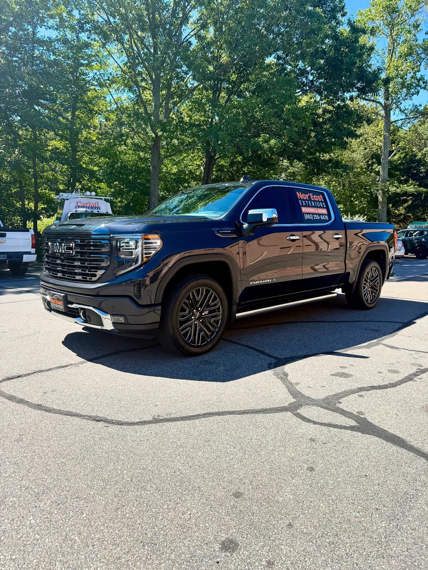 Dark blue GMC Sierra truck parked on pavement, trees in the background. Sunny day.