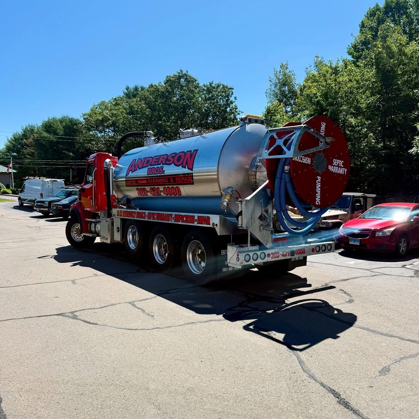 Red septic truck parked on asphalt, blue sky, trees in background.
