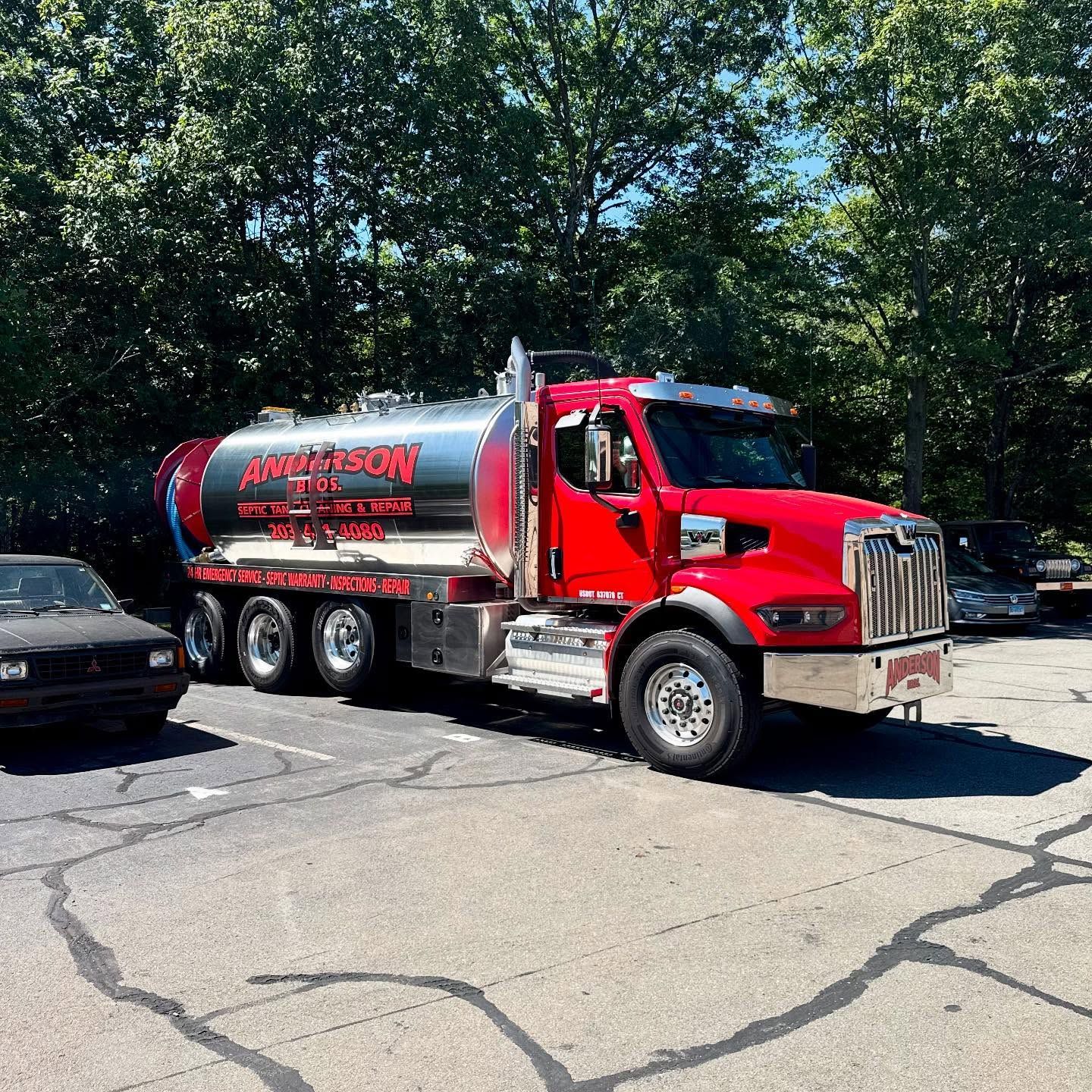 Red septic truck parked in a parking lot with a silver tank, trees in the background.