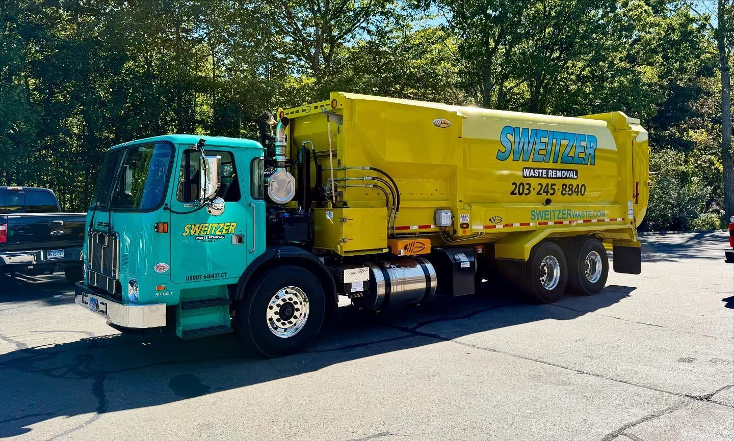 A teal and yellow garbage truck parked on pavement. The truck has a business name