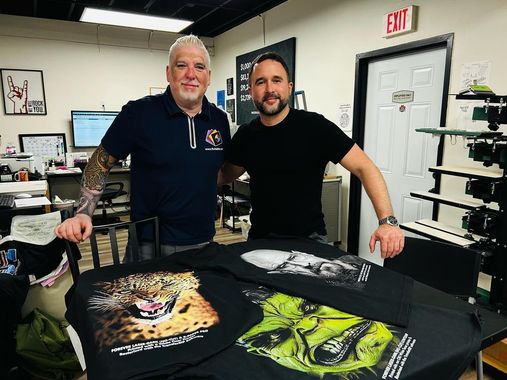 Two men stand near printed t-shirts in a print shop. One man has tattoos and the other has a beard.