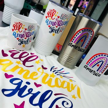 White shirt and drinkware with "All American Girl" and "America" graphics in patriotic colors.