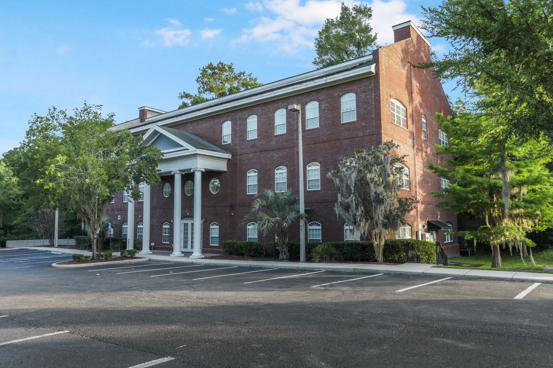 Brick building with white columns, windows, and trees, in a parking lot.