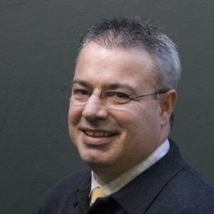 Man in glasses smiles, wearing a blazer with a yellow tie, in front of a dark background.