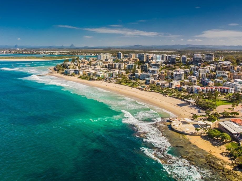 Aerial view of a coastal city with turquoise water, a sandy beach, and buildings under a blue sky.