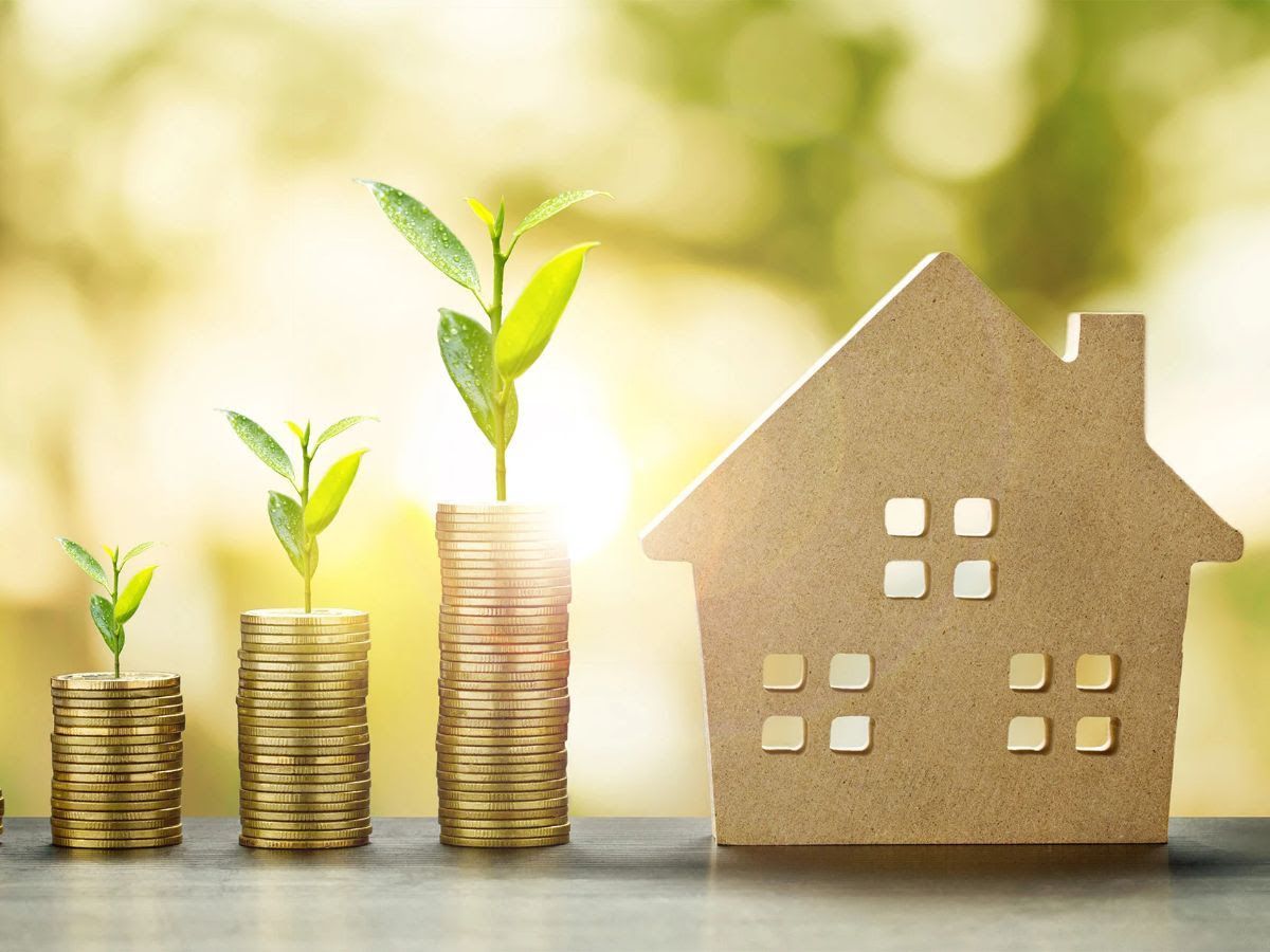 Piles of coins with growing plants, next to a wooden house cutout.