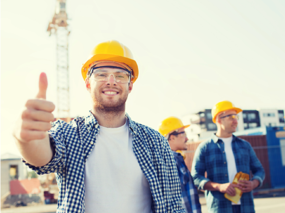 Construction worker giving a thumbs-up, smiling, wearing hard hat and safety glasses, with two colleagues in background.