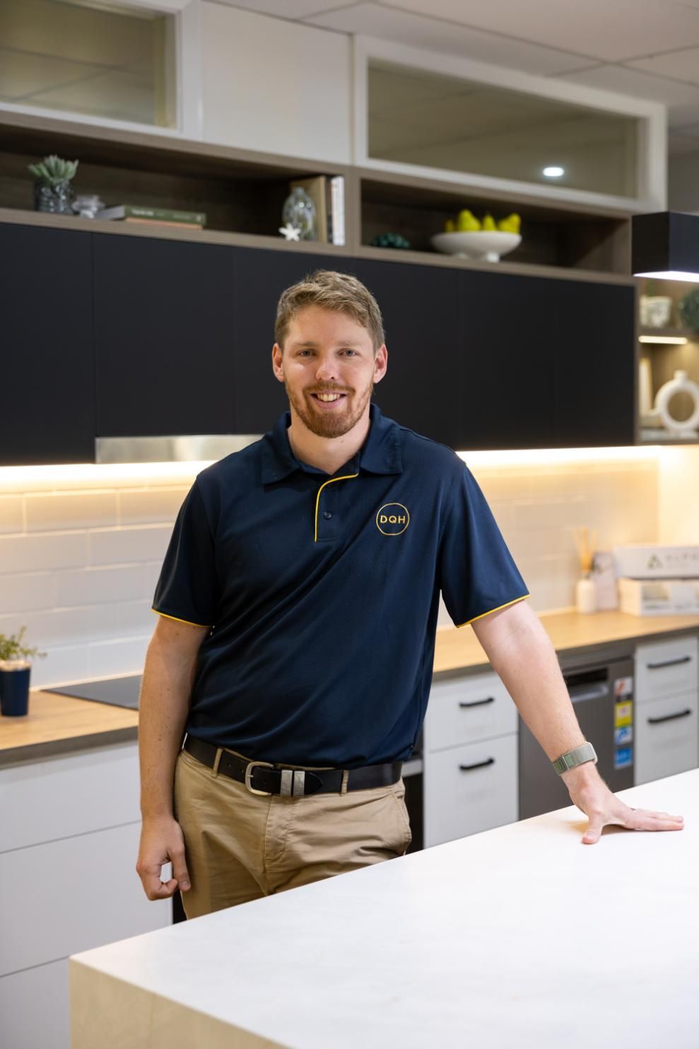 Man in navy polo shirt smiles, leaning on kitchen island, modern kitchen in background.