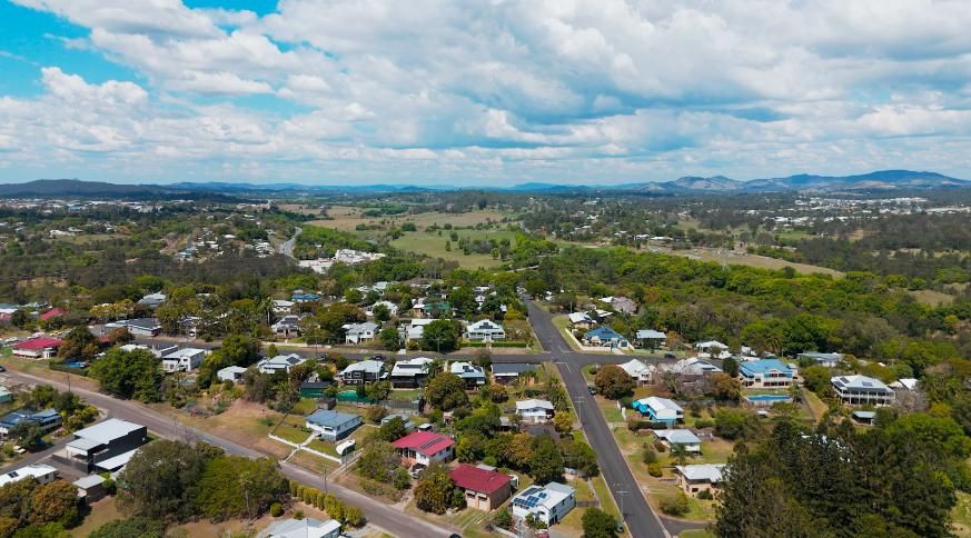 Aerial view of a suburban neighborhood with houses, streets, trees, and a distant green field under a cloudy sky.