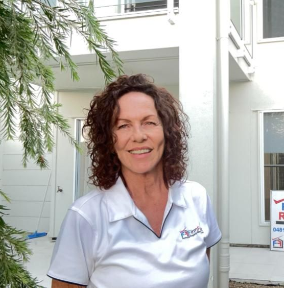 Woman with curly hair smiles while using a laptop in a light-filled room with white shelves.