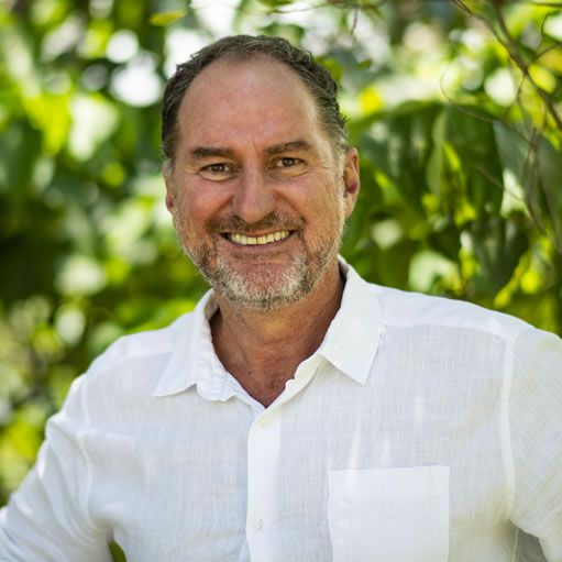 Man with short hair and a beard smiling, wearing a white shirt, set against a green, leafy background.