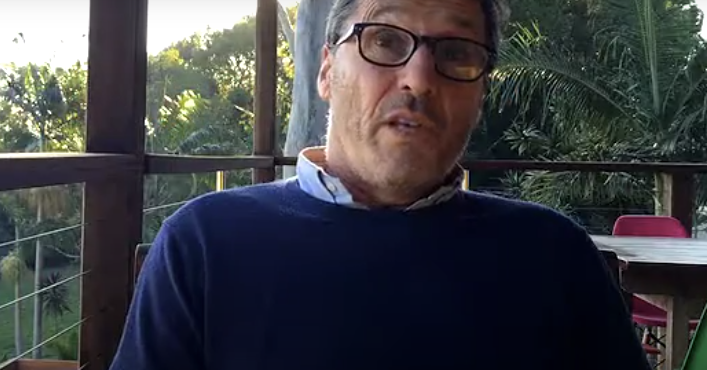 Man wearing glasses and blue sweater, speaking outdoors on a porch.