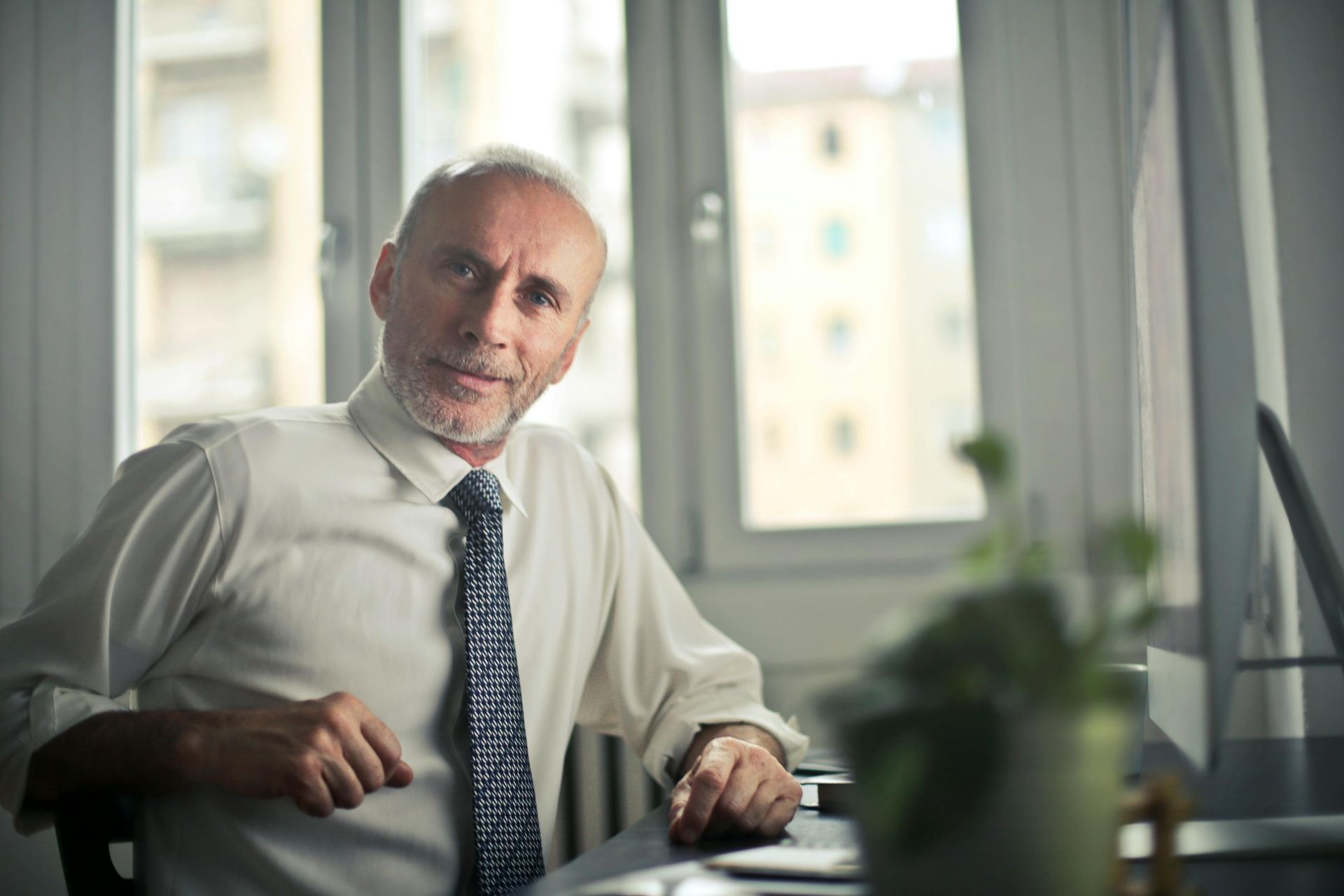 Man in a button-down shirt and tie sits at a desk, looking towards the camera with a neutral expression.