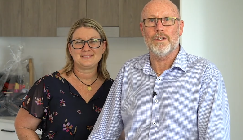 Woman and man standing together indoors. Man wears glasses and blue striped shirt. Woman wears glasses and floral top.