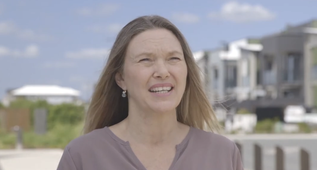 Woman with long hair speaking outdoors in front of modern houses.
