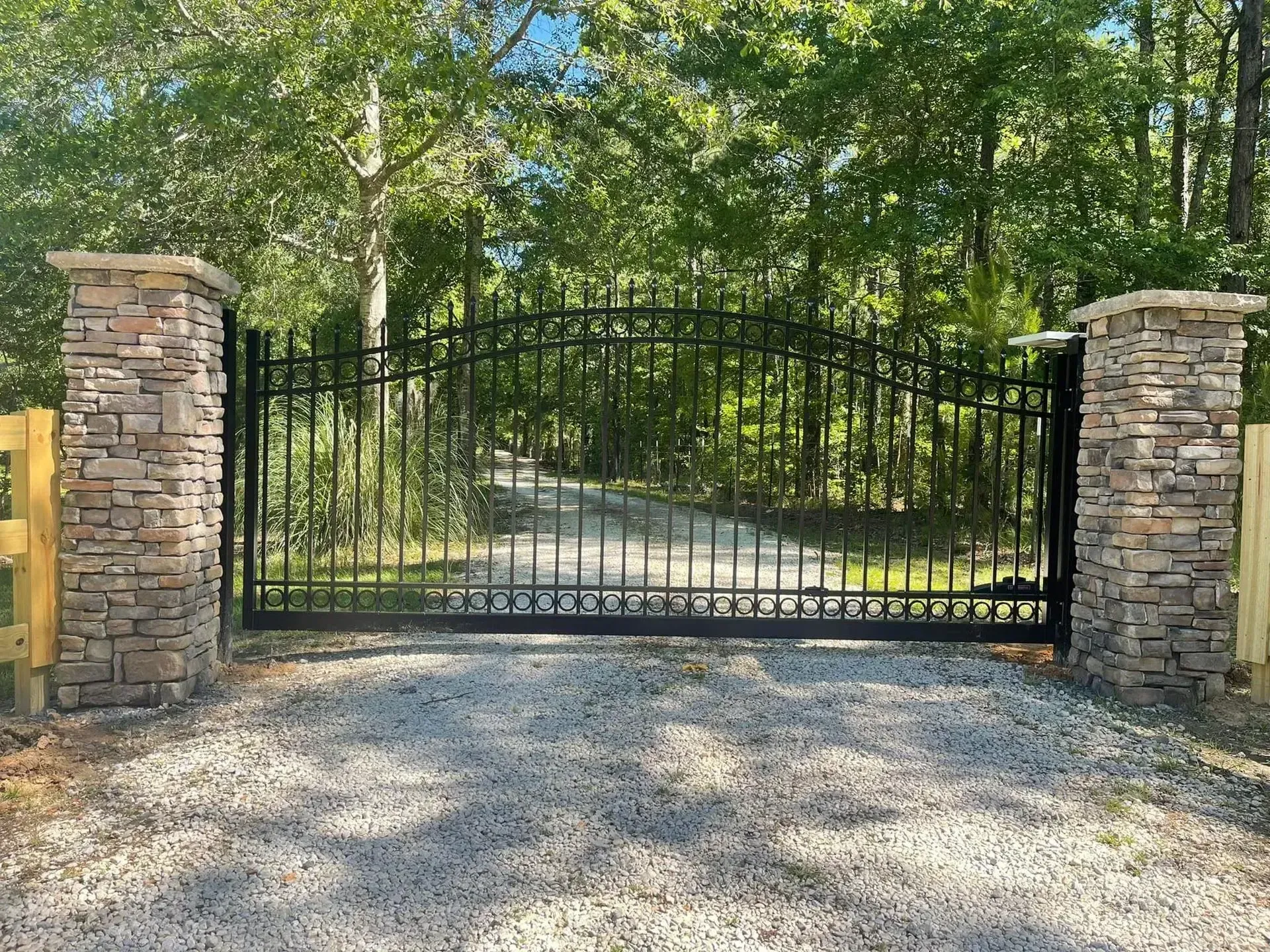 A metal gate is surrounded by stone pillars on the side of a gravel road.