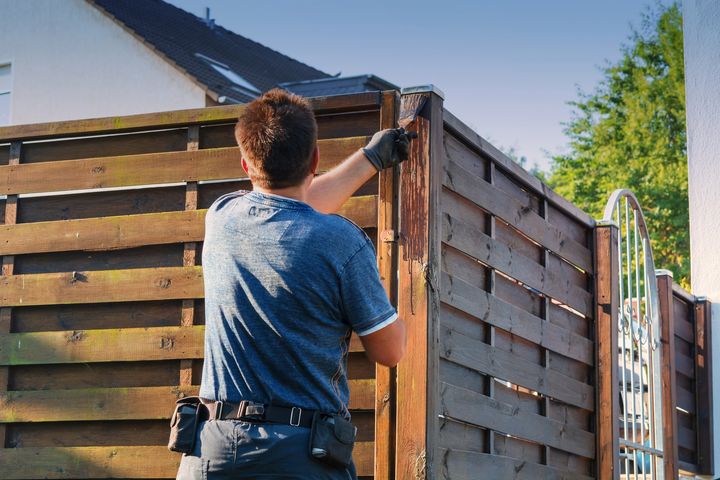 A man is painting a wooden fence in front of a house.