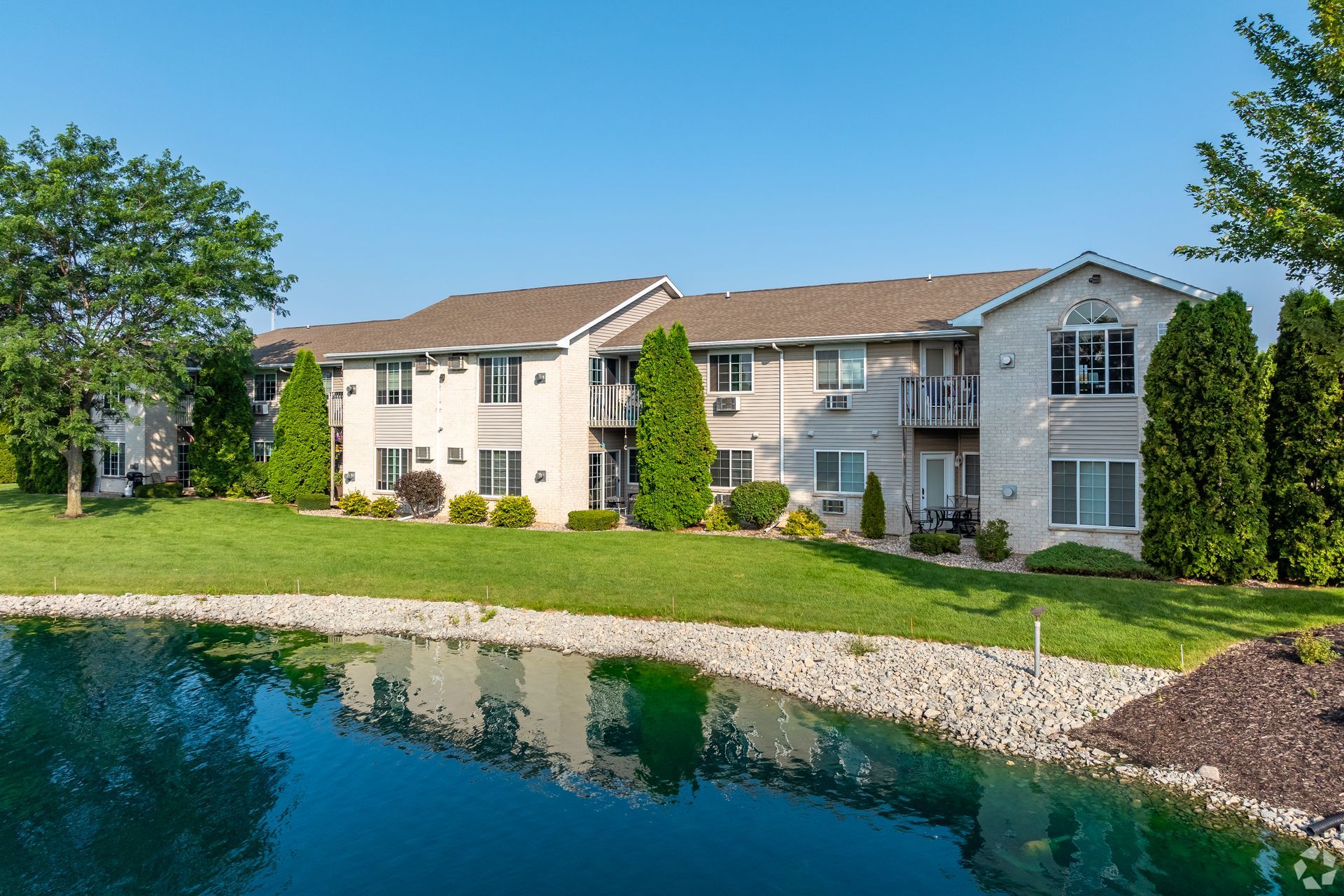 Apartment buildings beside a pond with green lawn and trees on a sunny day