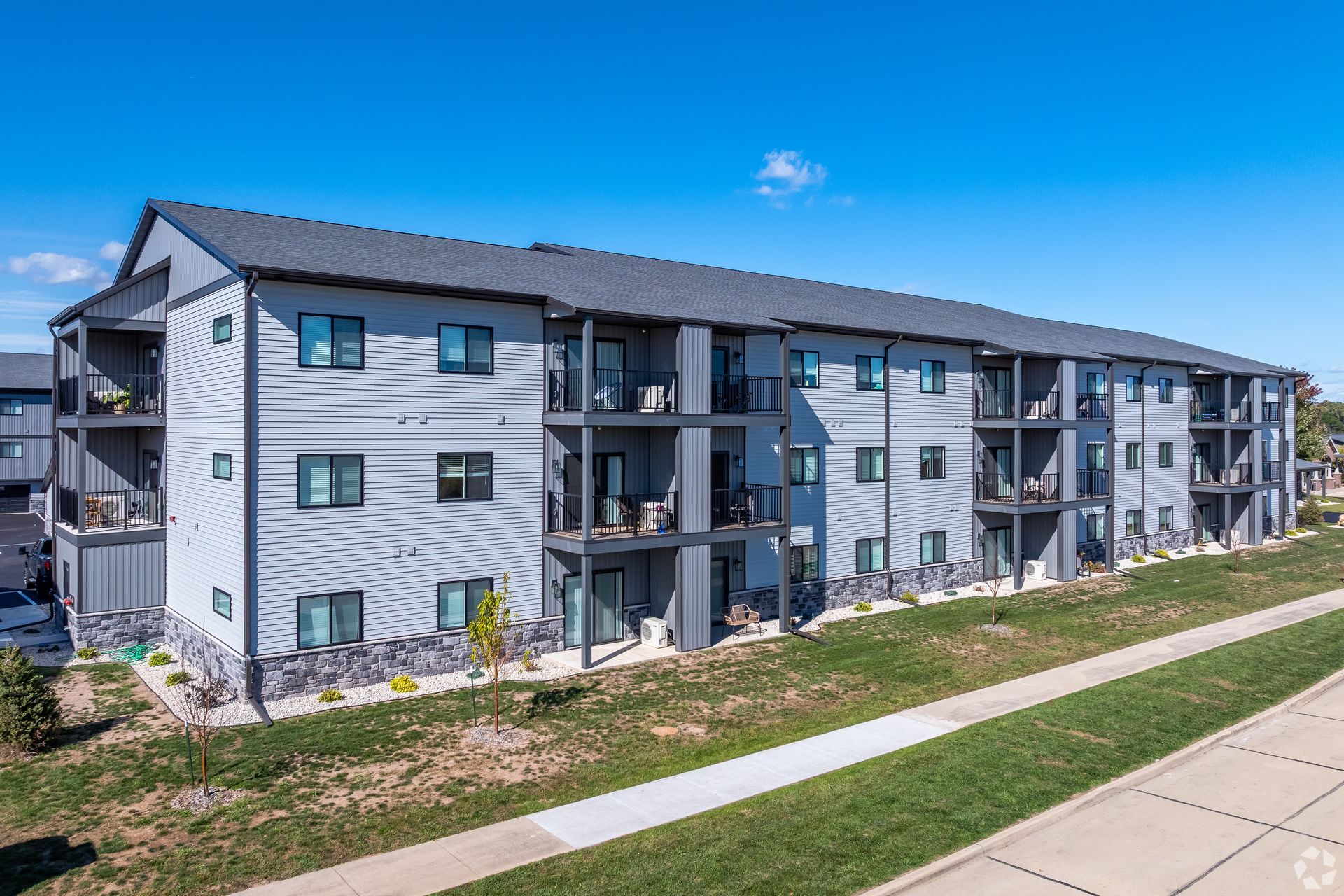 Three-story gray apartment building with balconies beside a sidewalk under a blue sky