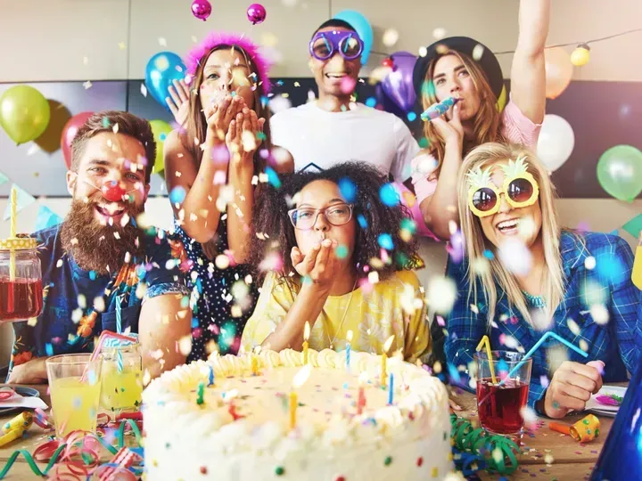 Friends celebrating a birthday, blowing confetti. Cake with candles, balloons, festive accessories.