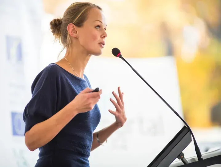 Woman giving a presentation at a podium, gesturing with her hands; soft background.