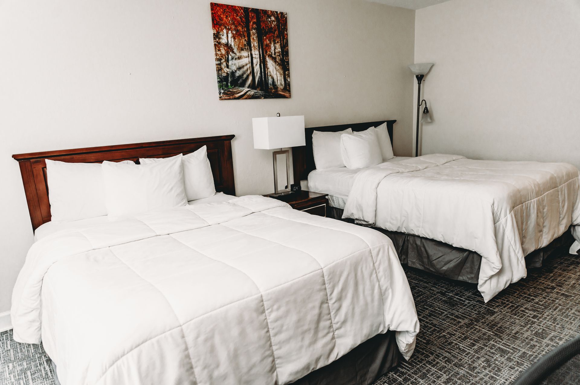 Two beds with white bedding in a hotel room with a painting and floor lamp.