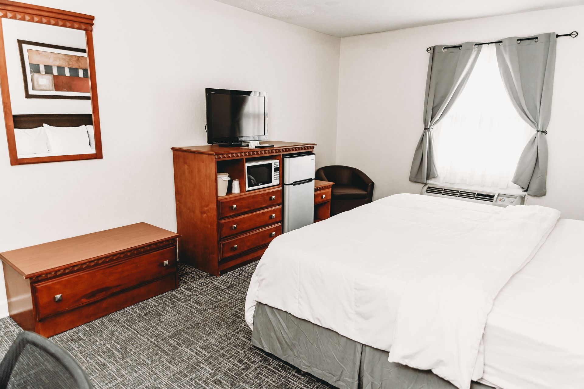 Hotel room with bed, dresser, TV, mini-fridge, window, and armchair.