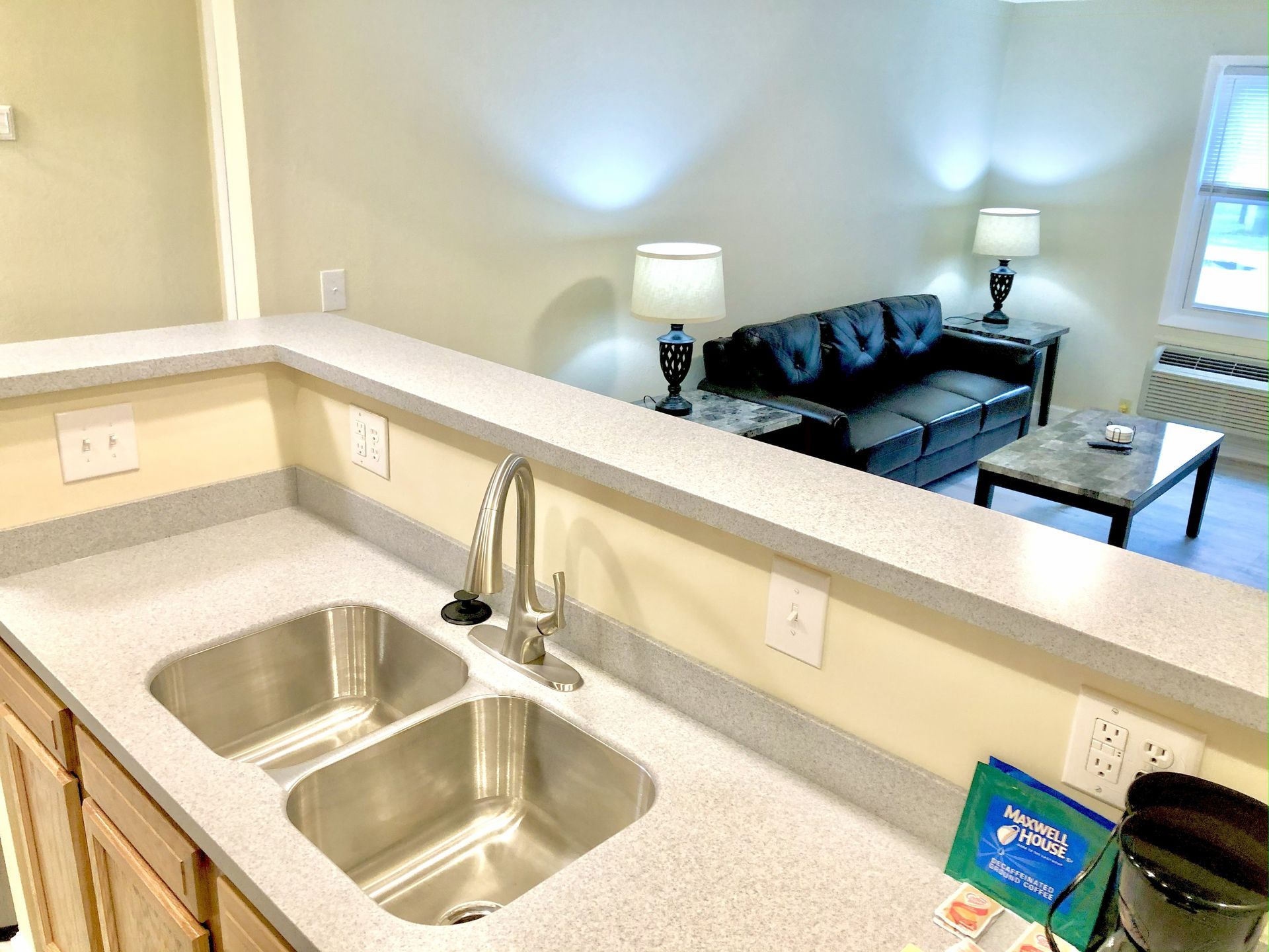 Kitchen with stainless steel sink, countertop, and a living area with a couch and lamps in the background.
