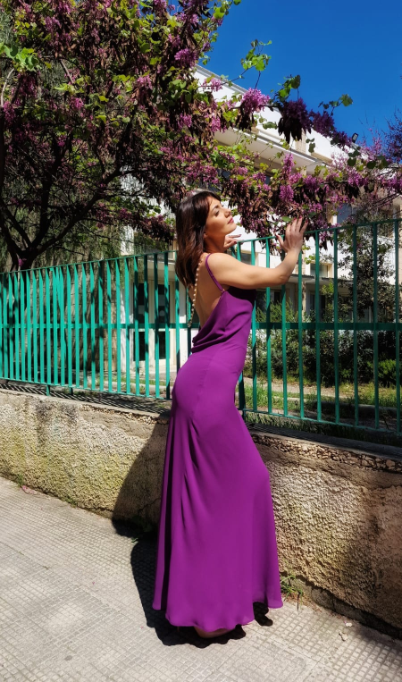 Una donna con un abito viola posa accanto a una recinzione verde, sotto fiori in fiore e un cielo azzurro.