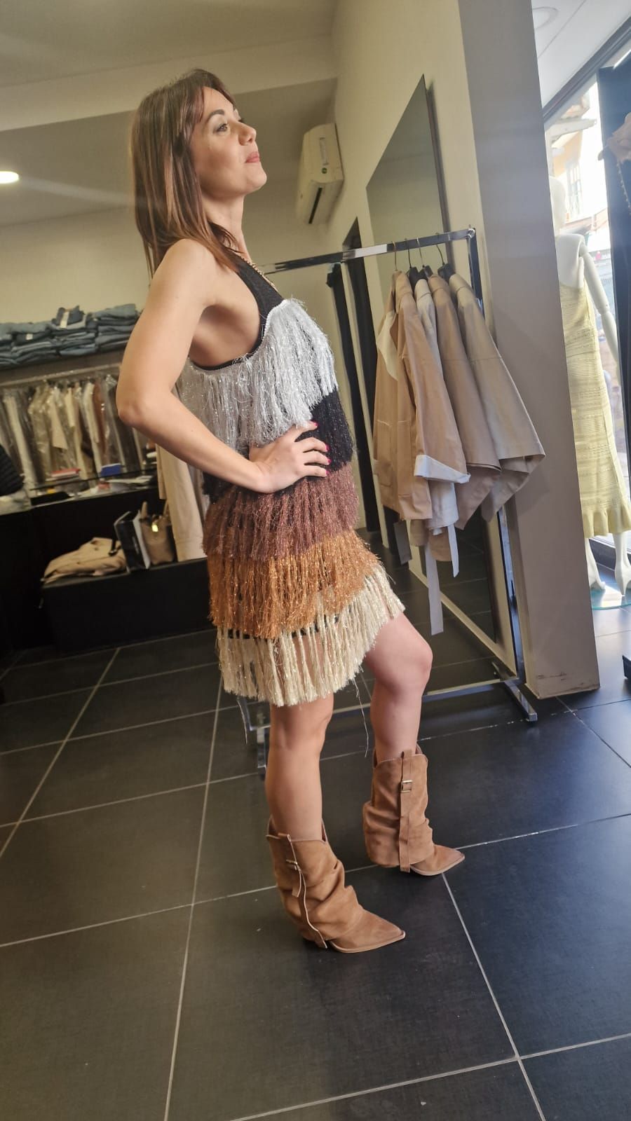 Una persona posa con un abito boho con frange e stivali color cuoio all'interno di una boutique di abbigliamento.