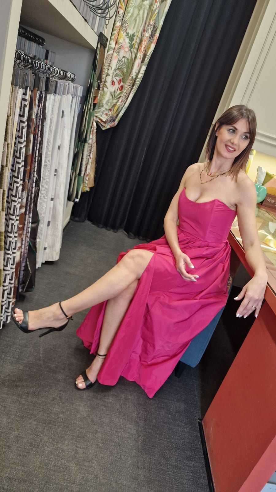 Una donna in abito rosa è seduta in un camerino, sorride con una gamba accavallata.