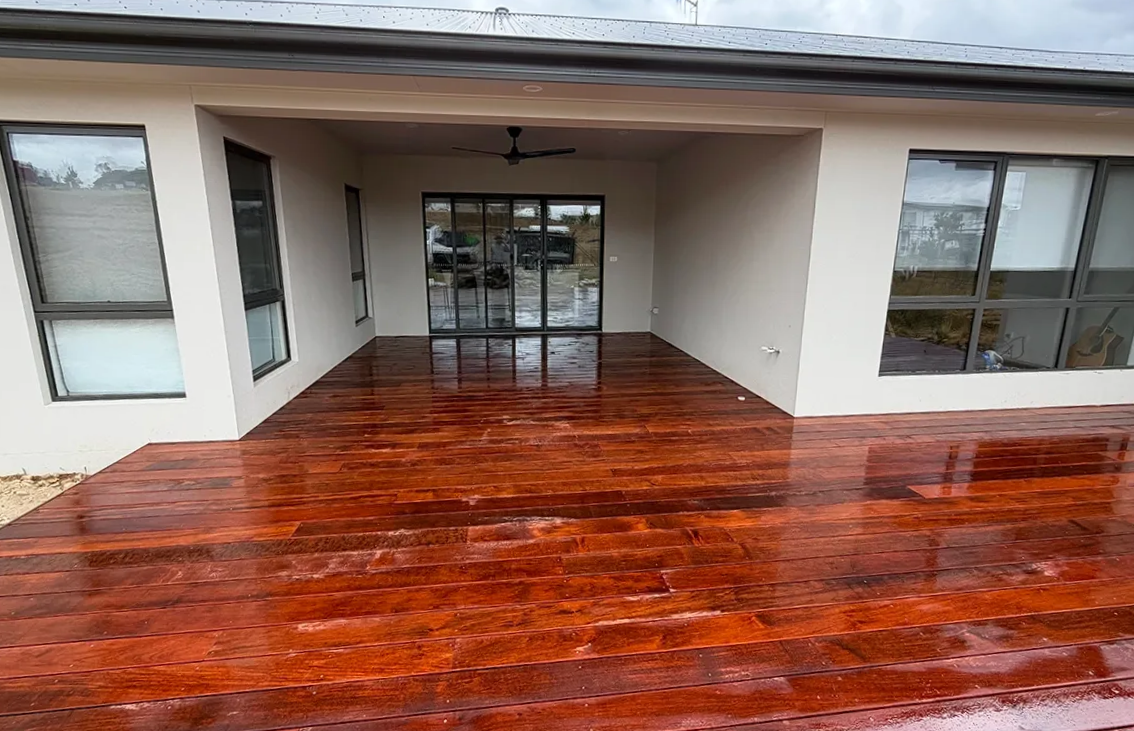 Covered wooden deck with a glossy red finish, next to a house with large windows. — Straight up Building Projects in Holt, ACT