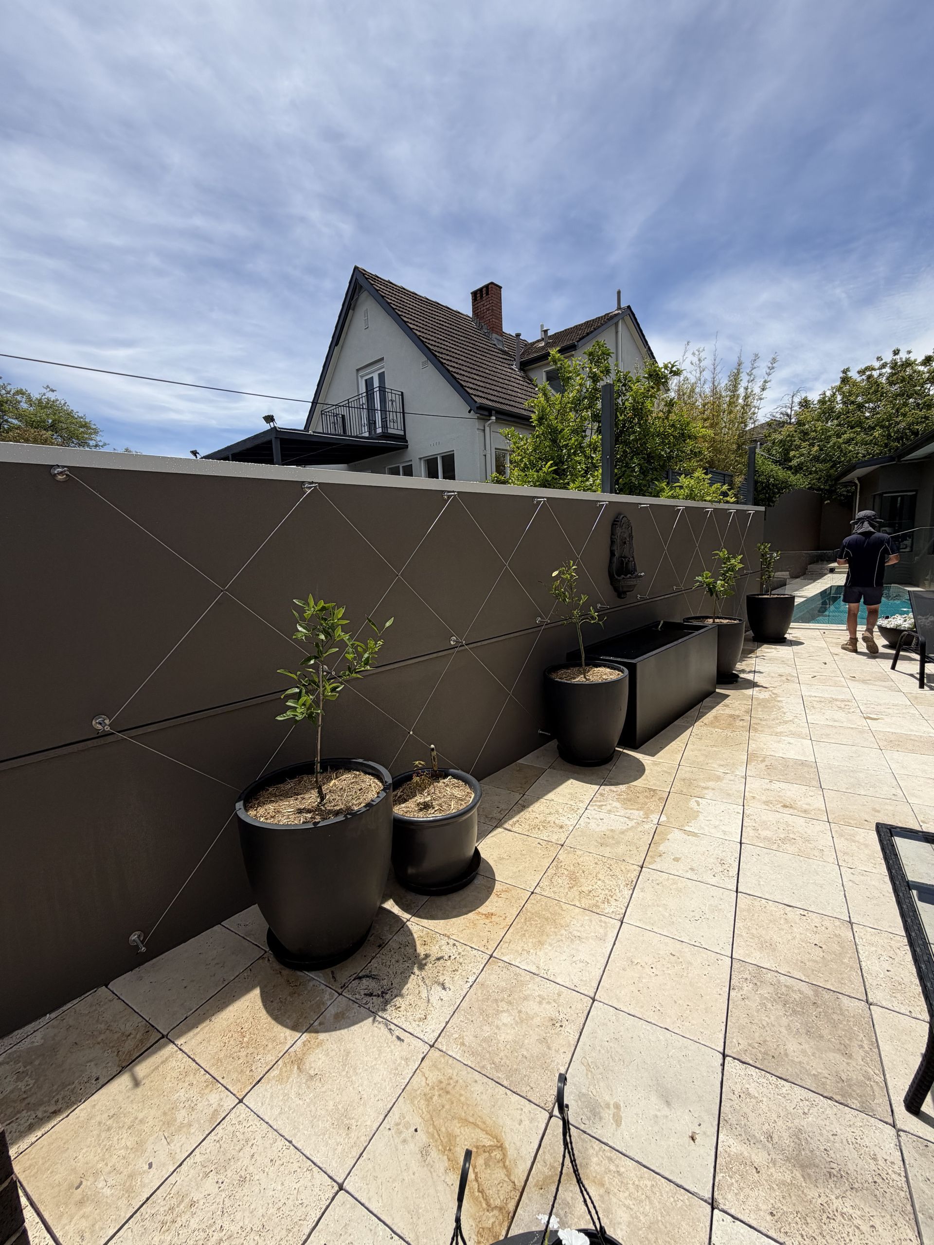 A patio with travertine tiles, a dark wall featuring diamond-patterned wire, and several potted plants near a pool — Straight up Building Projects in Ainslie, ACT