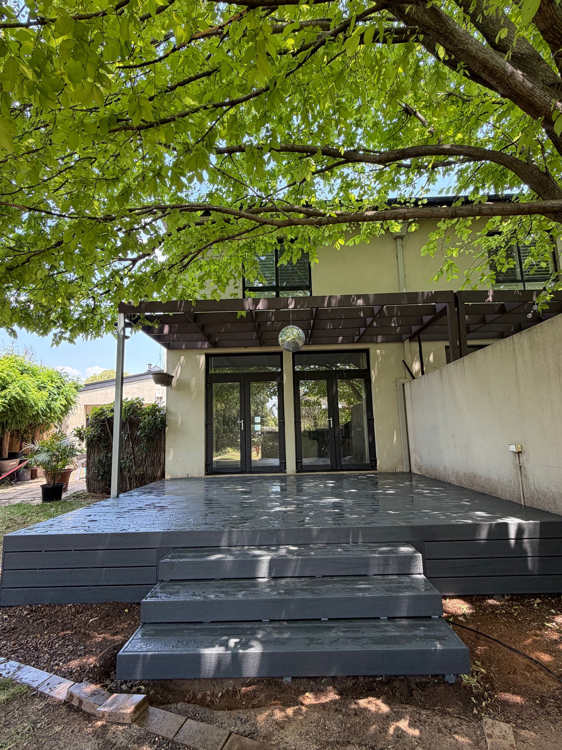 Gray deck with steps leading to glass doors, under a leafy tree. Beige building in background. — Straight up Building Projects in Holt, ACT