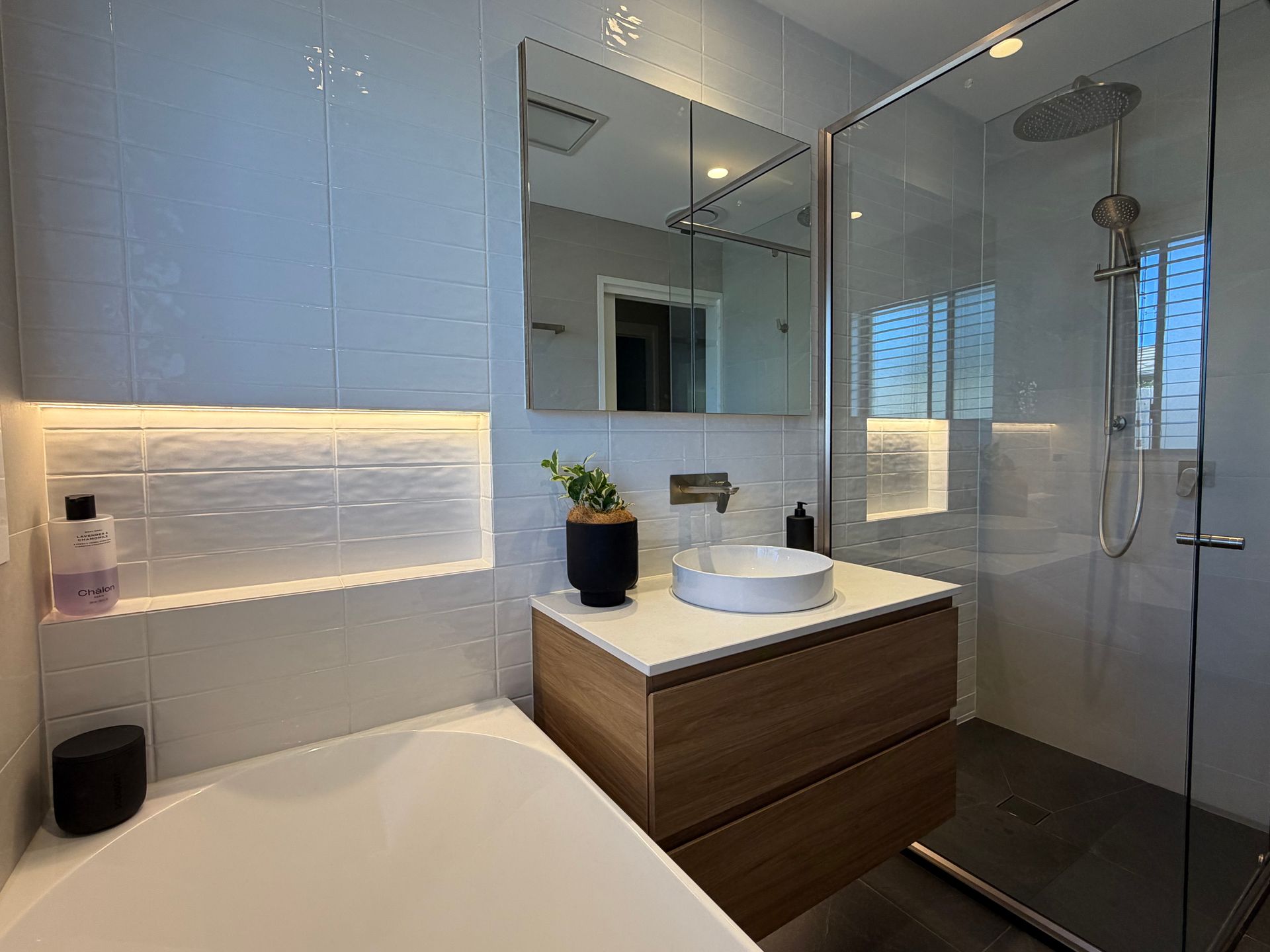 Modern bathroom with a wooden vanity, a white sink, a glass shower, and a built-in shelf with lighting. — Straight up Building Projects in Holt, ACT