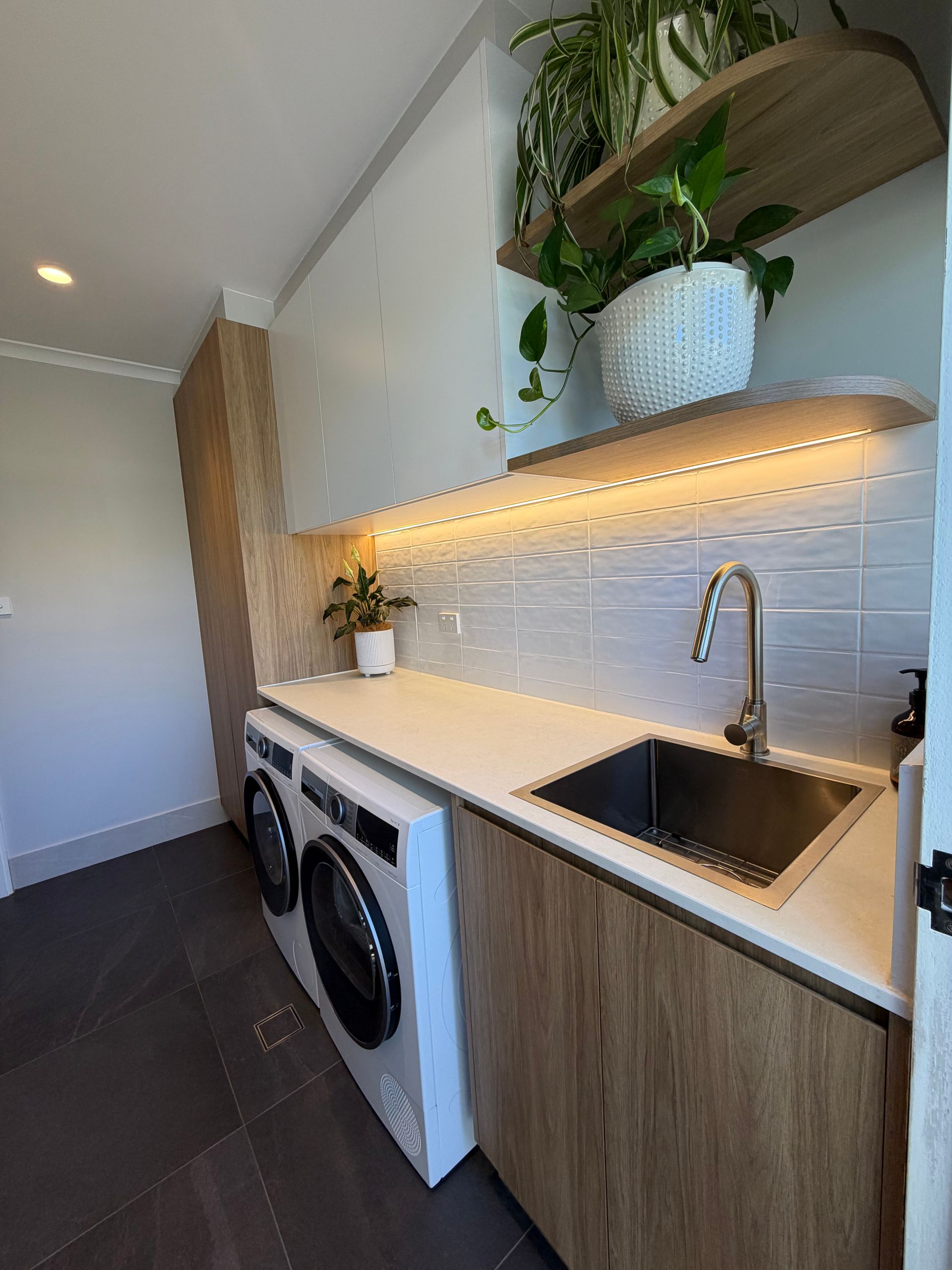Laundry room with washer, dryer, sink, wooden cabinets, white countertop, and shelves with plants. — Straight up Building Projects in Holt, ACT