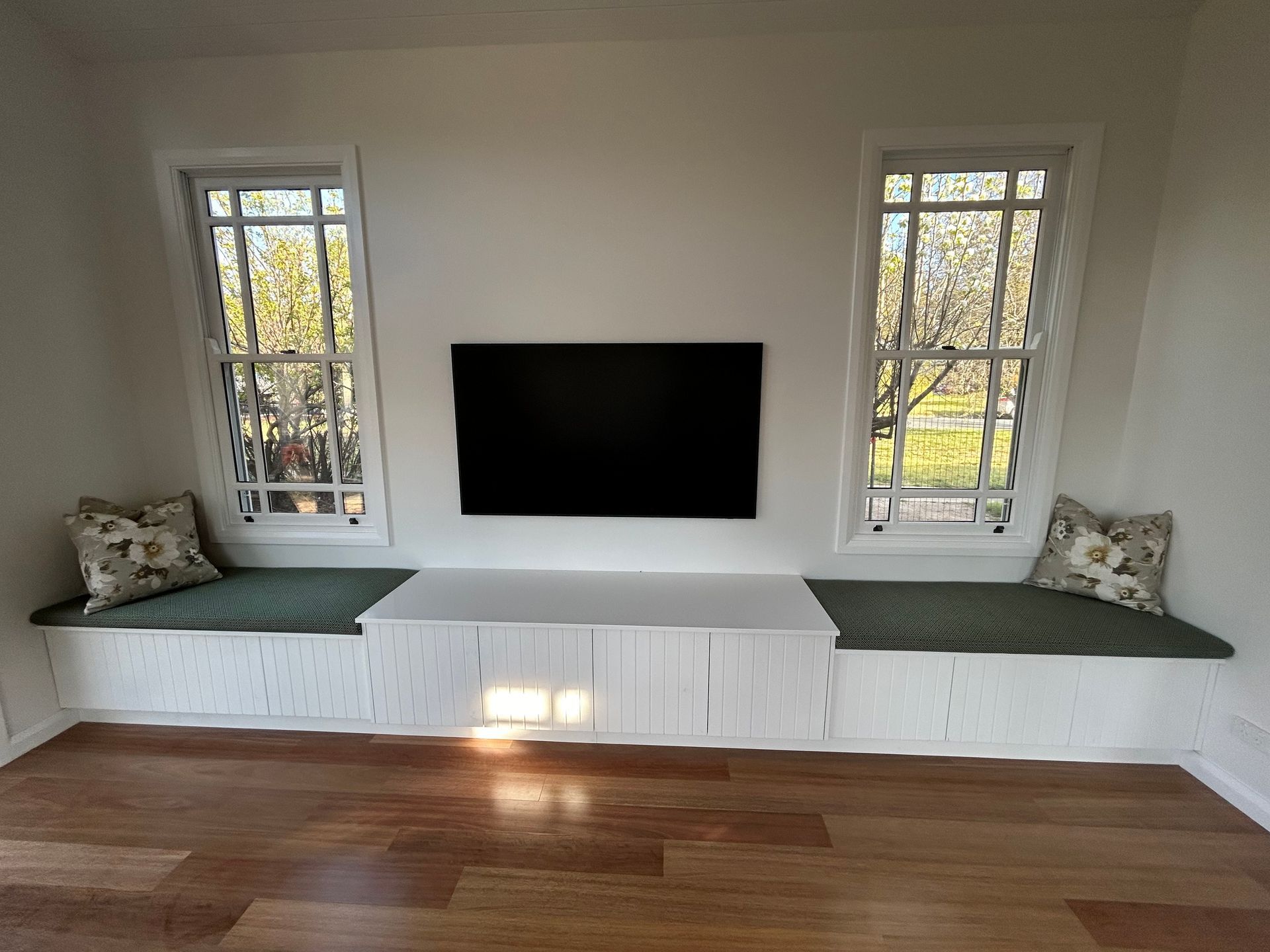 Built-in white entertainment center with a mounted TV, flanked by two windows. Includes a seating bench with green cushions and pillows. — Straight up Building Projects in Holt, ACT