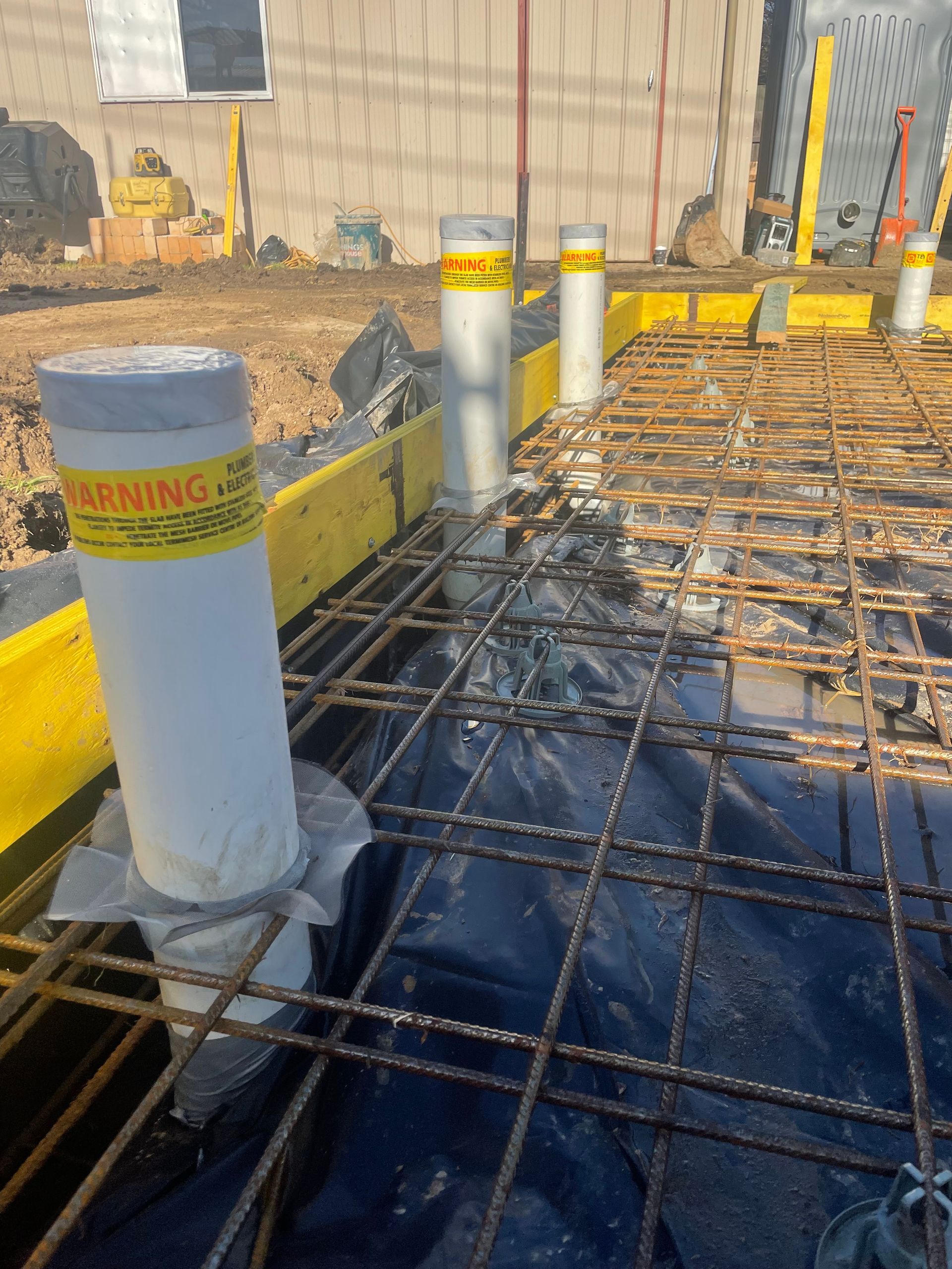 Construction site: White pipes with warning labels protrude from rebar and plastic sheeting, next to yellow formwork. — Straight up Building Projects in Holt, ACT