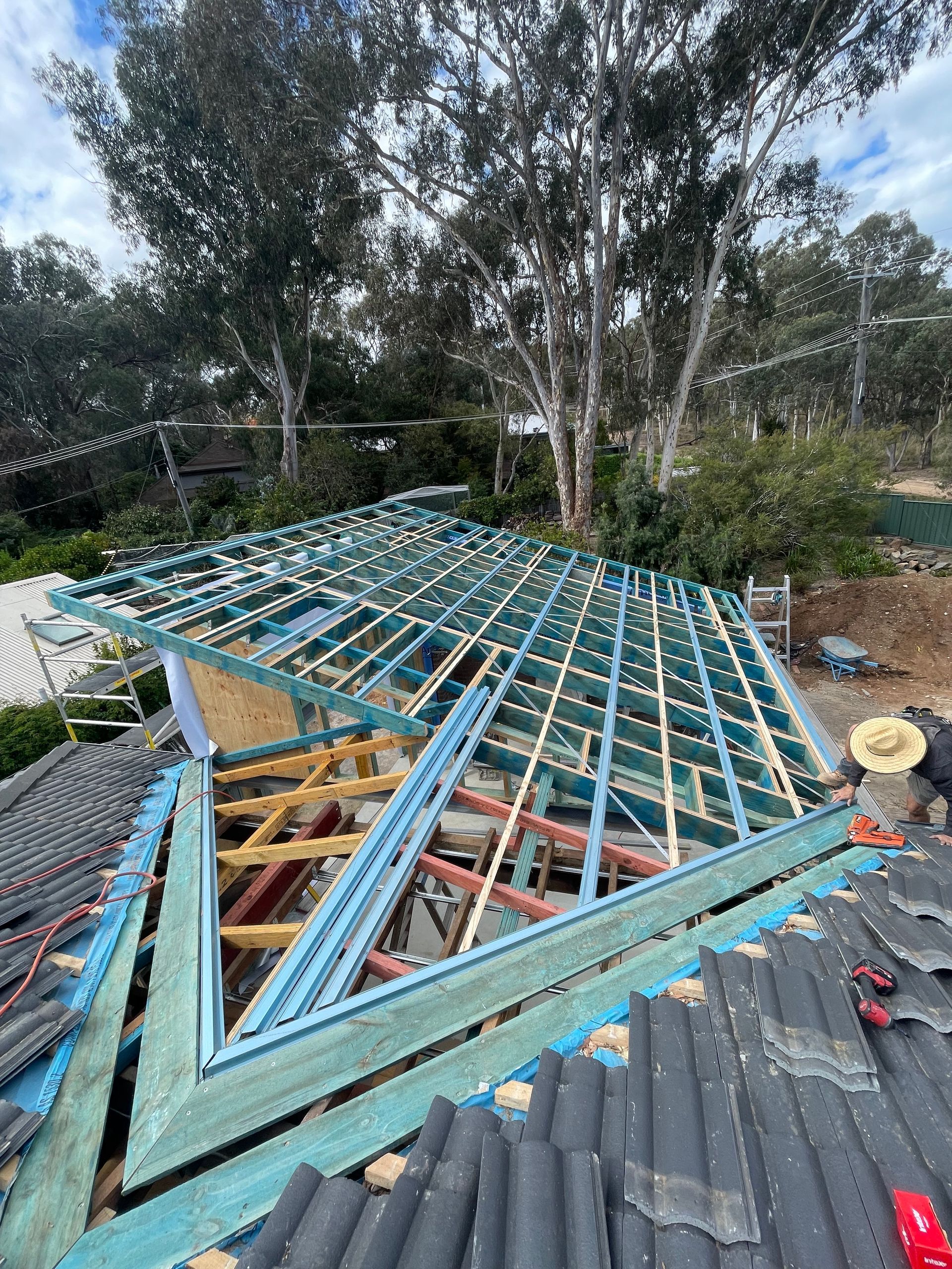 A construction worker on a residential roof installing a wooden frame for a roof extension against a backdrop of trees.
