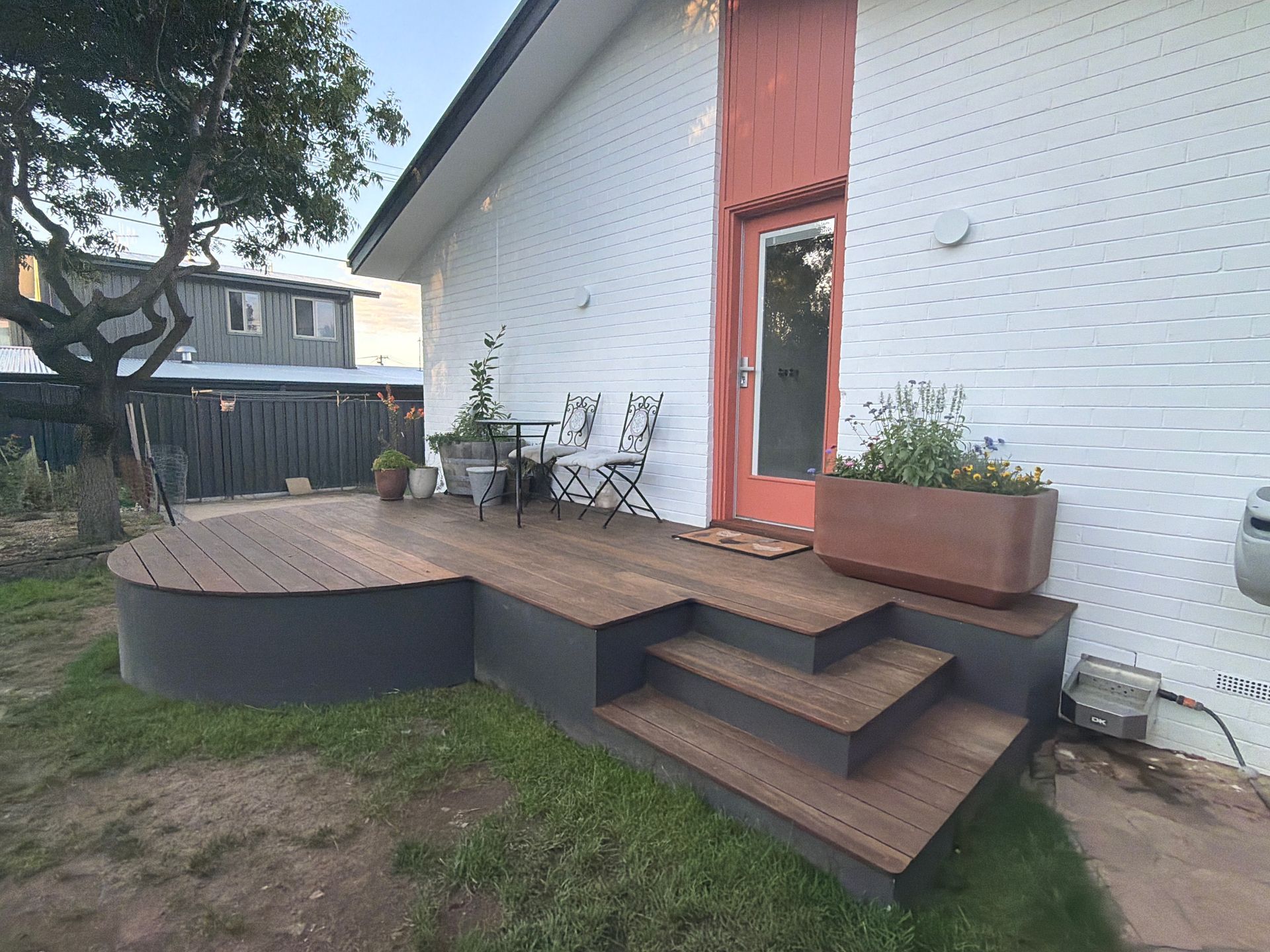 A modern wooden deck with a curved edge and stairs, attached to a white house with a coral-coloured door frame  — Straight up Building Projects in Holt, ACT