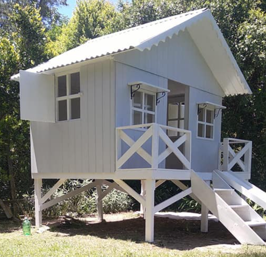 Casa de juegos elevada de color blanco con porche y escalones, ubicada en un patio con césped.