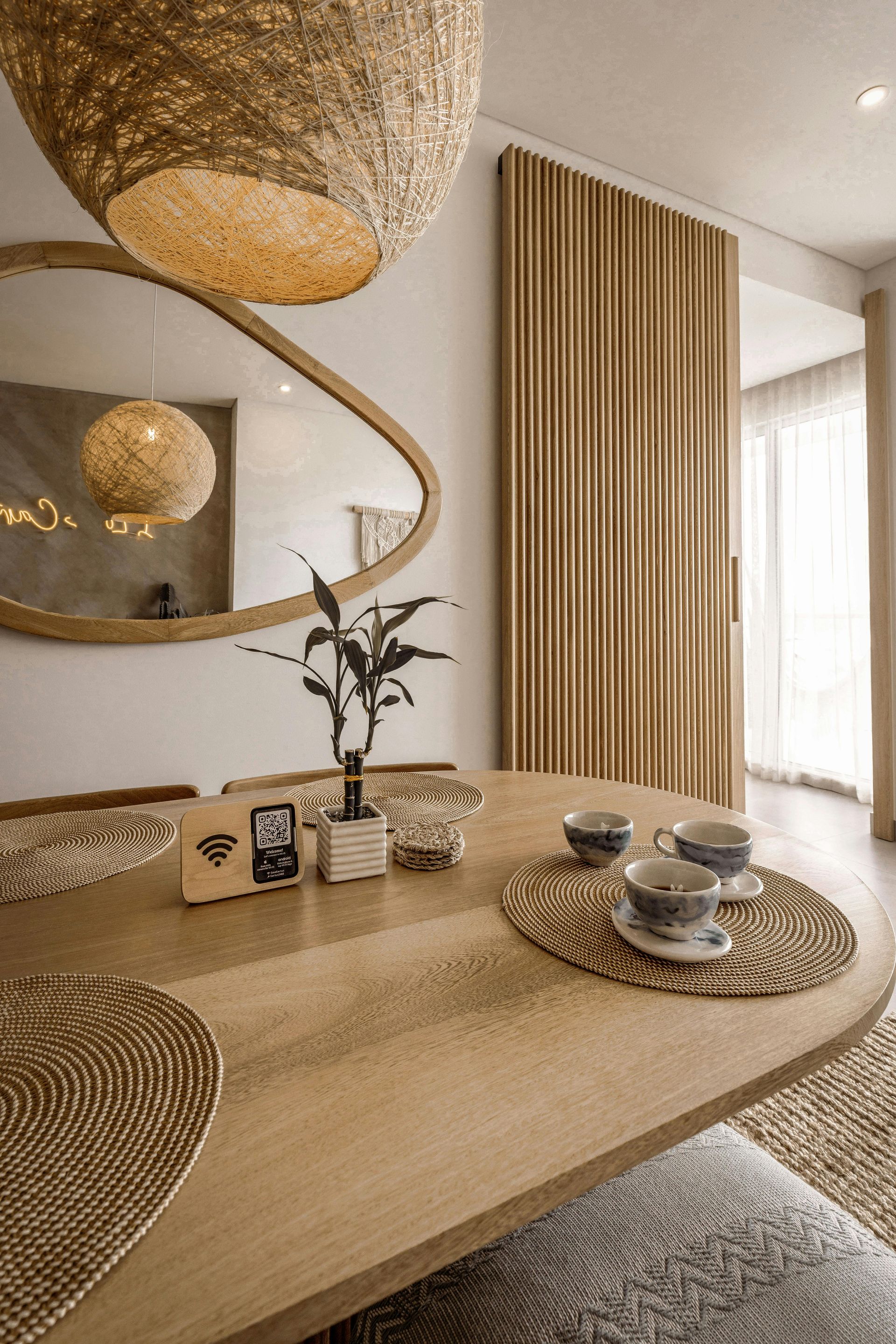 Comedor con mesa de madera, espejo redondo, lámpara tejida y pared decorativa de madera.