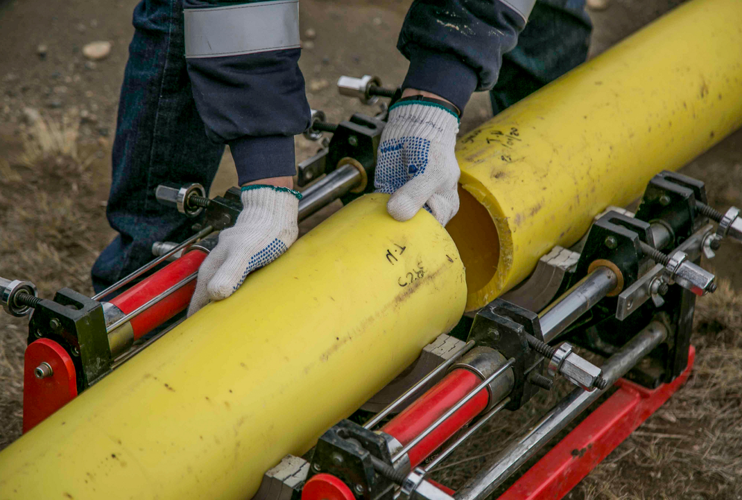 Worker's gloved hands positioning a yellow pipe on machinery rollers outdoors