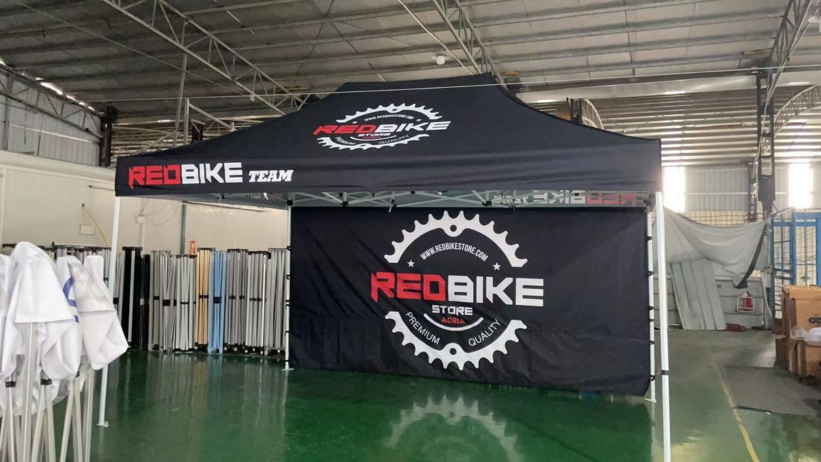 RedBike motorcycle display booth with black canopy, logo banner, and hanging white apparel in an indoor hall