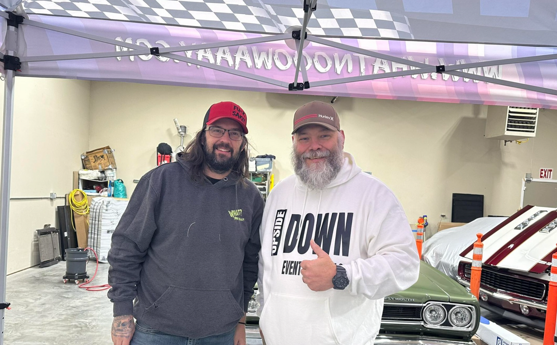 Two men posing under a canopy at a car show, one in a red cap and one giving a thumbs-up.