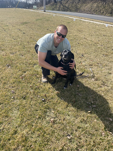 A man is kneeling down next to a black dog in the grass.