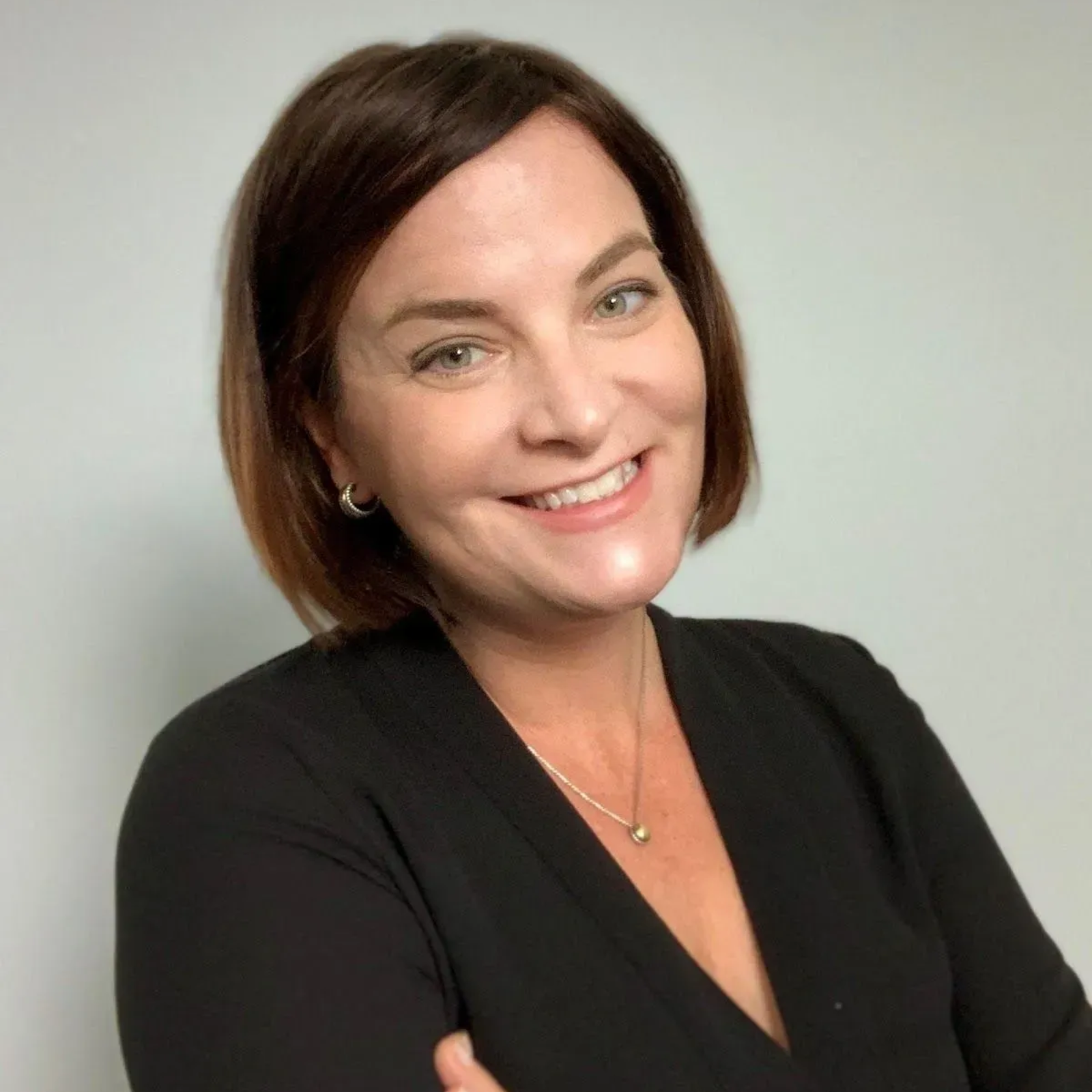 Woman with brown hair smiles, wearing a black top and a silver necklace, indoors.